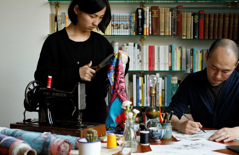 A female artist cutting fabric with scissors while a male artist draws a sketch with a pencil.